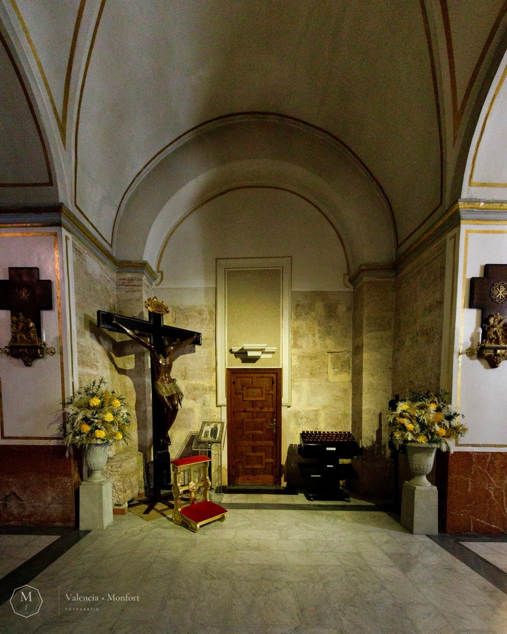 Capilla con un crucificado entre dos jarrones de flores amarillas, junto a una puerta de madera